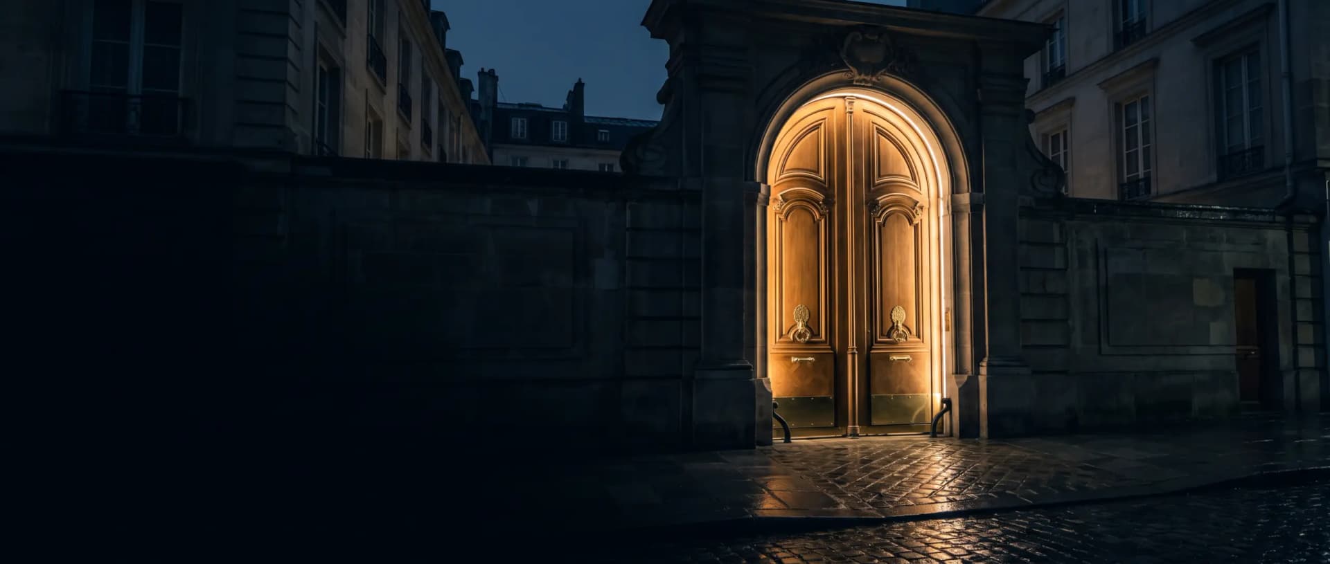 Porte cochère en pierre à clef de voûte éclairée de l'intérieur, pavés mouillés, nuit bleue